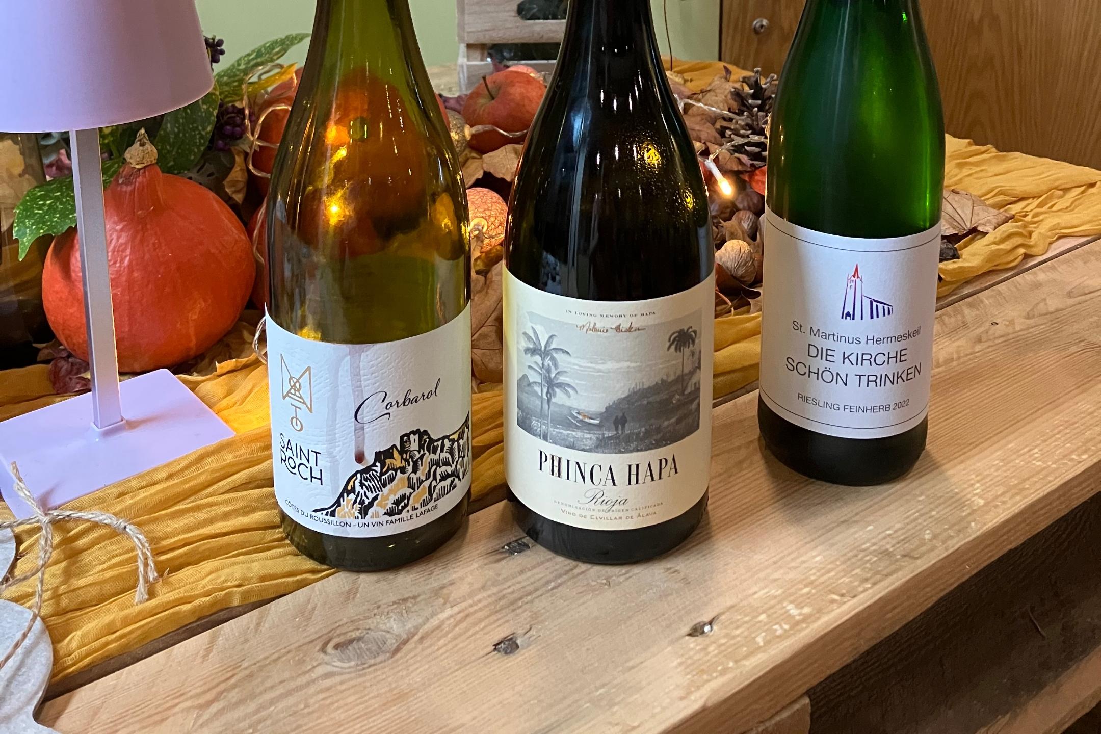 Drei Flaschen mit verschiedenen Weinen auf einem Holztisch. Die Weine tragen die Namen von Heiligen. Im Hintergrund: Herbstliche Dekoration.